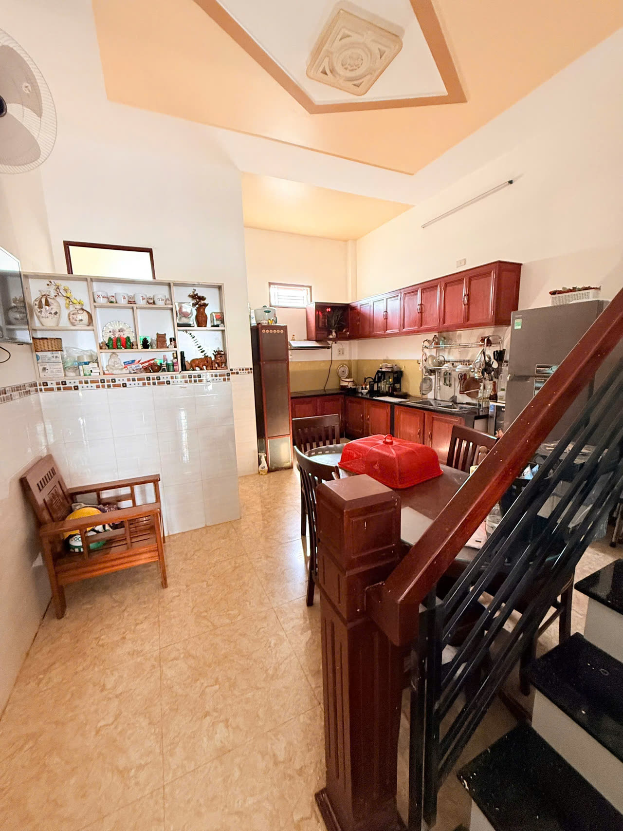 Khu bếp nhà Diệp Minh Tuyền Nha Trang với tủ bếp gỗ đỏ, bàn ăn và tủ lạnh, cạnh chân cầu thang