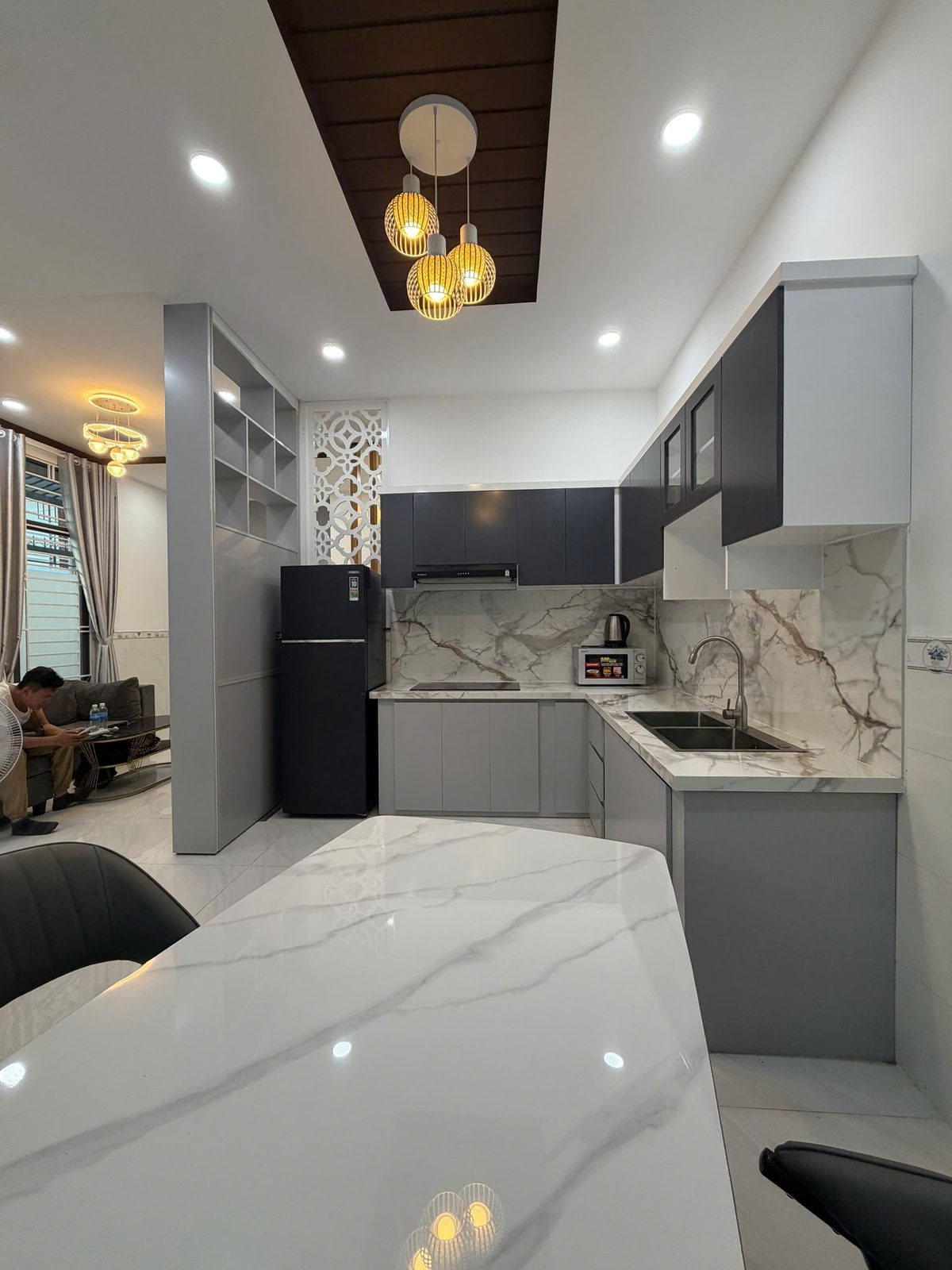 Bếp chữ L nhà Vĩnh Điềm với tủ bếp hai tông xám trắng, mặt đá vân mây, bếp từ, máy hút mùi và bàn đảo mặt đá phía trước