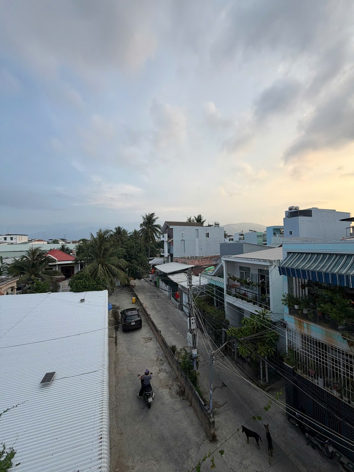 Đường bê tông trước nhà Vĩnh Điềm nhìn từ ban công trên cao, có một ô tô màu đen đậu sát lề và một xe máy đang chạy qua