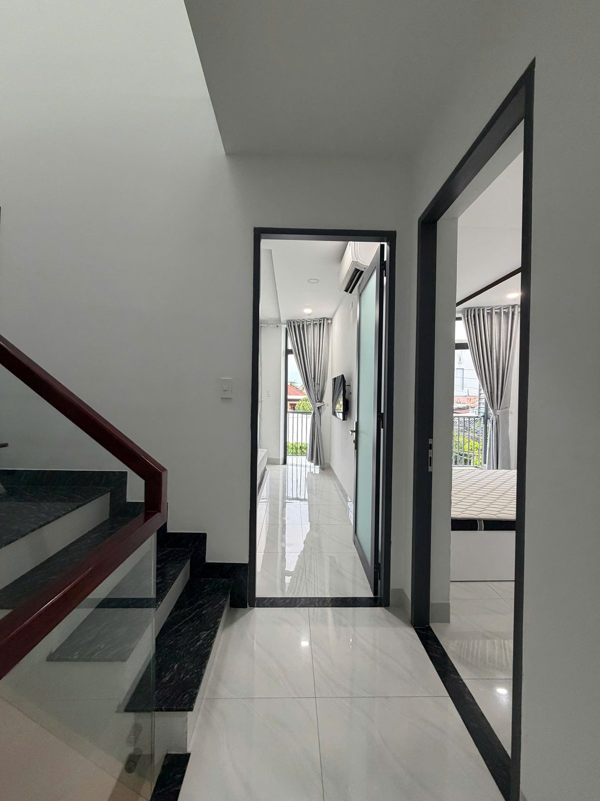 Hành lang tầng trên nhà Vĩnh Điềm với cửa kính khung nhôm đen dẫn vào hai phòng ngủ, nền gạch bóng màu trắng