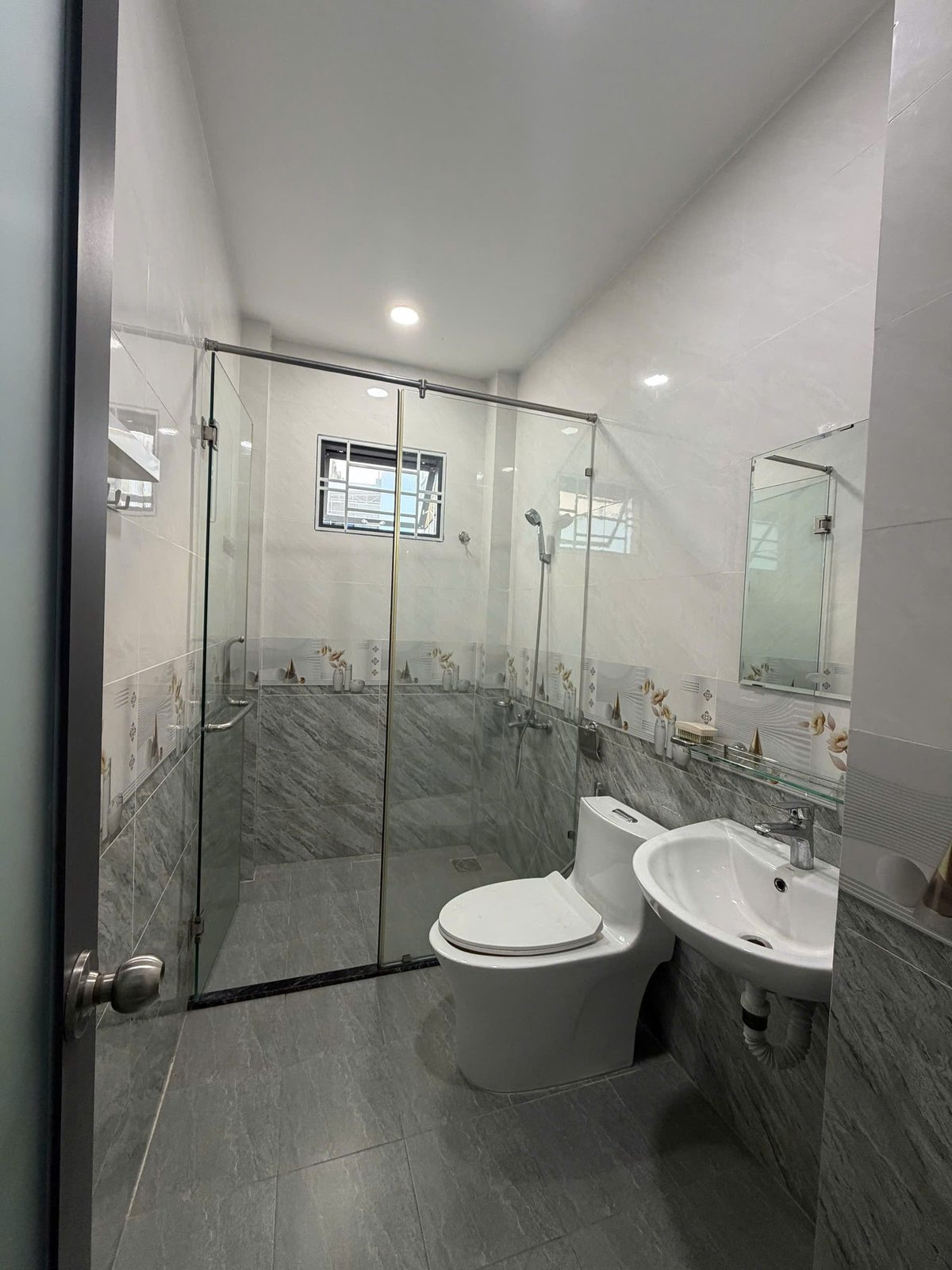 Nhà vệ sinh nhà Vĩnh Điềm ốp gạch giả đá xám, có vách kính tách khu tắm, sen tắm, bồn cầu, lavabo và gương treo tường