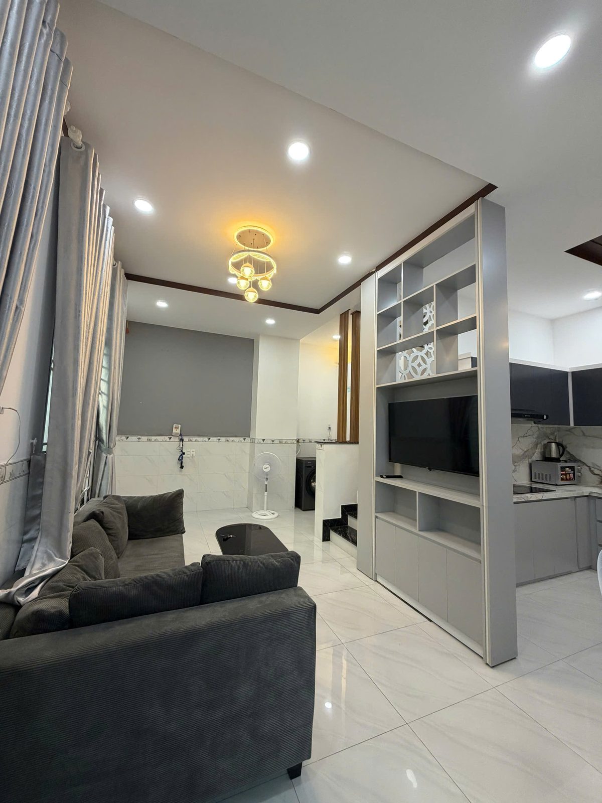 Phòng khách nhà Vĩnh Điềm với sofa xám, kệ tivi làm vách ngăn với khu bếp phía sau, trần thạch cao có đèn âm