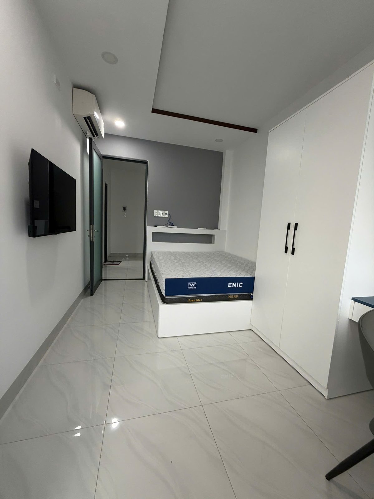 Phòng ngủ nhỏ nhà Vĩnh Điềm với giường đơn bục liền, tủ áo trắng cao kịch trần, bàn làm việc nhỏ và tivi treo tường