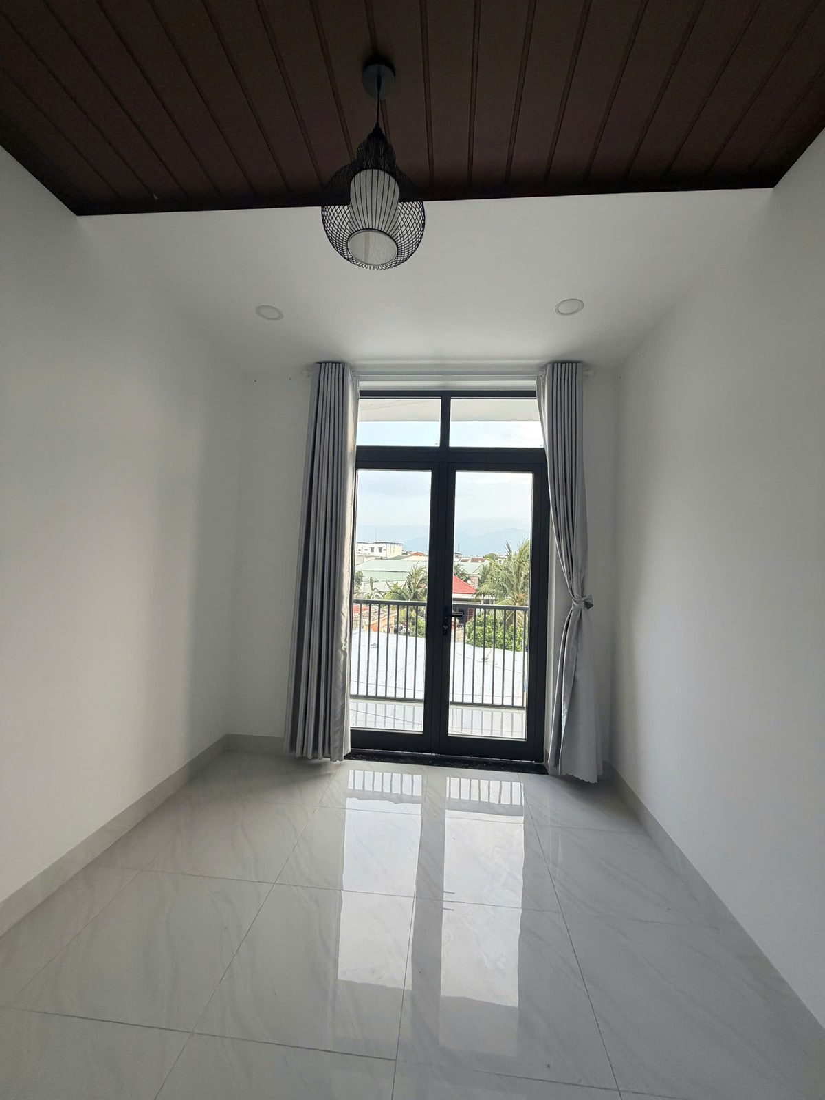 Phòng trống nhà Vĩnh Điềm với trần ốp gỗ màu nâu, đèn thả giữa phòng, nền gạch bóng và cửa kính mở ra ban công riêng