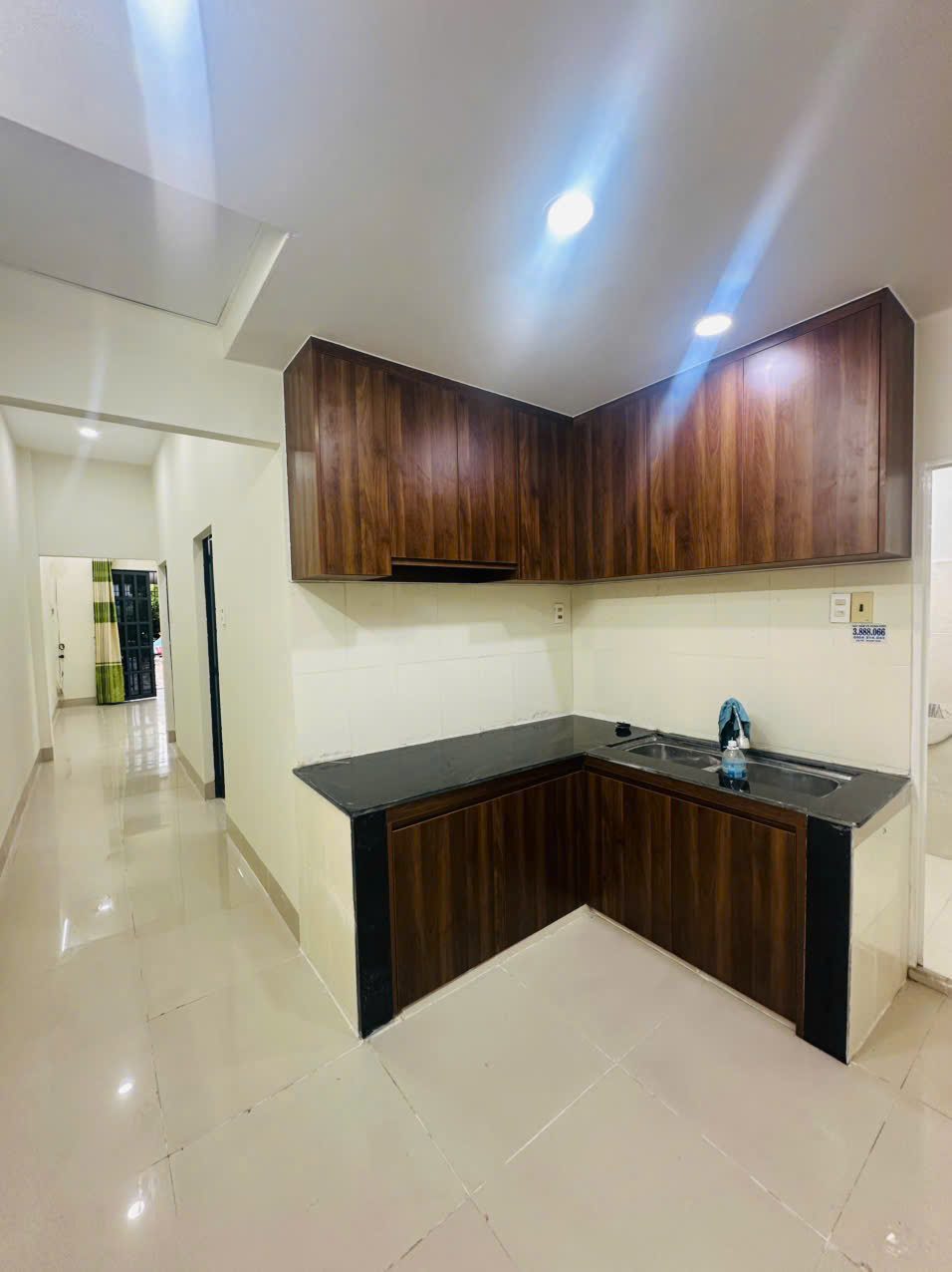 Bếp chữ L với tủ bếp gỗ màu nâu, mặt bàn đá đen và bồn rửa đôi, nhìn từ hành lang dẫn ra phòng khách