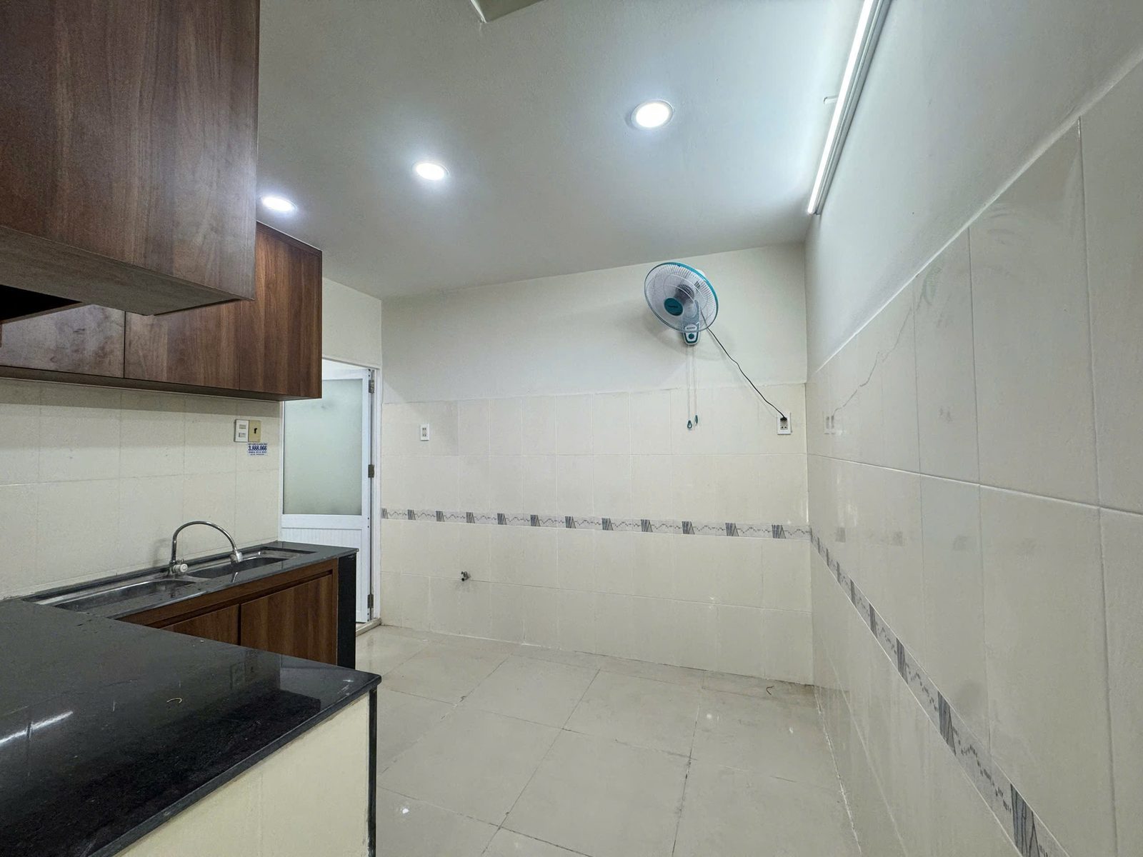 Khu bếp nhìn dọc theo tường ốp gạch men, có quạt treo tường và cửa kính mờ dẫn ra phía sau nhà