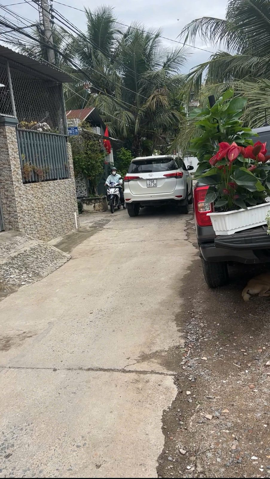 Hẻm bê tông trước nhà Vĩnh Ngọc với một xe bảy chỗ màu trắng và một xe bán tải đang đậu, xe máy vẫn đi qua được