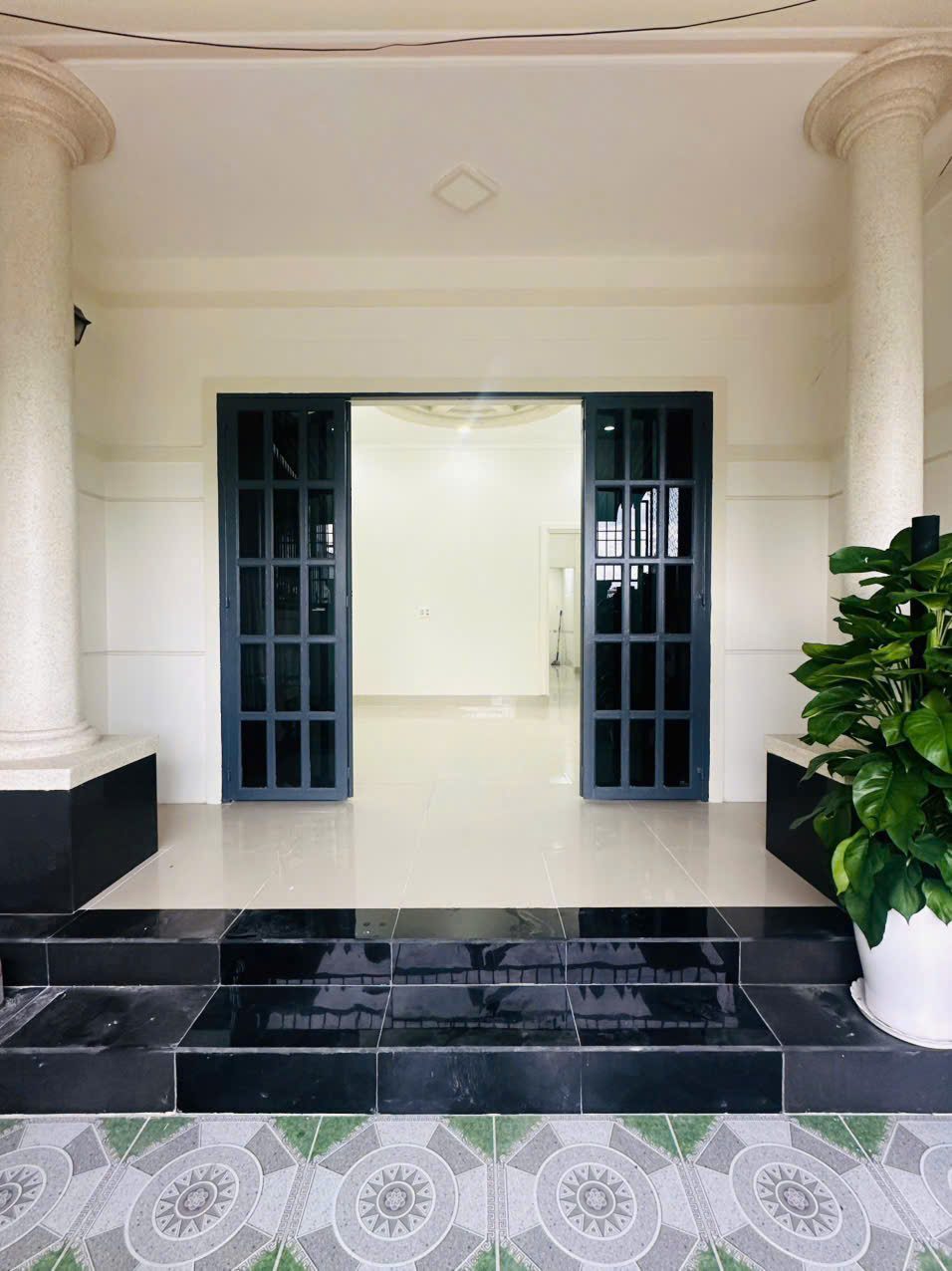 Hiên nhà với hai cột tròn, bậc tam cấp ốp đá đen và cửa chính gỗ kính mở vào phòng khách