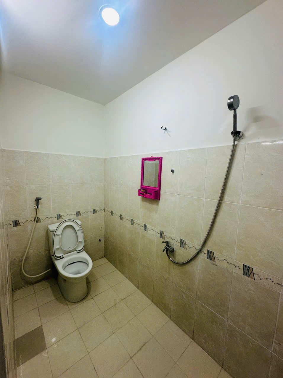 Nhà vệ sinh ốp gạch men màu kem, có bồn cầu, vòi sen tắm và gương nhỏ treo tường