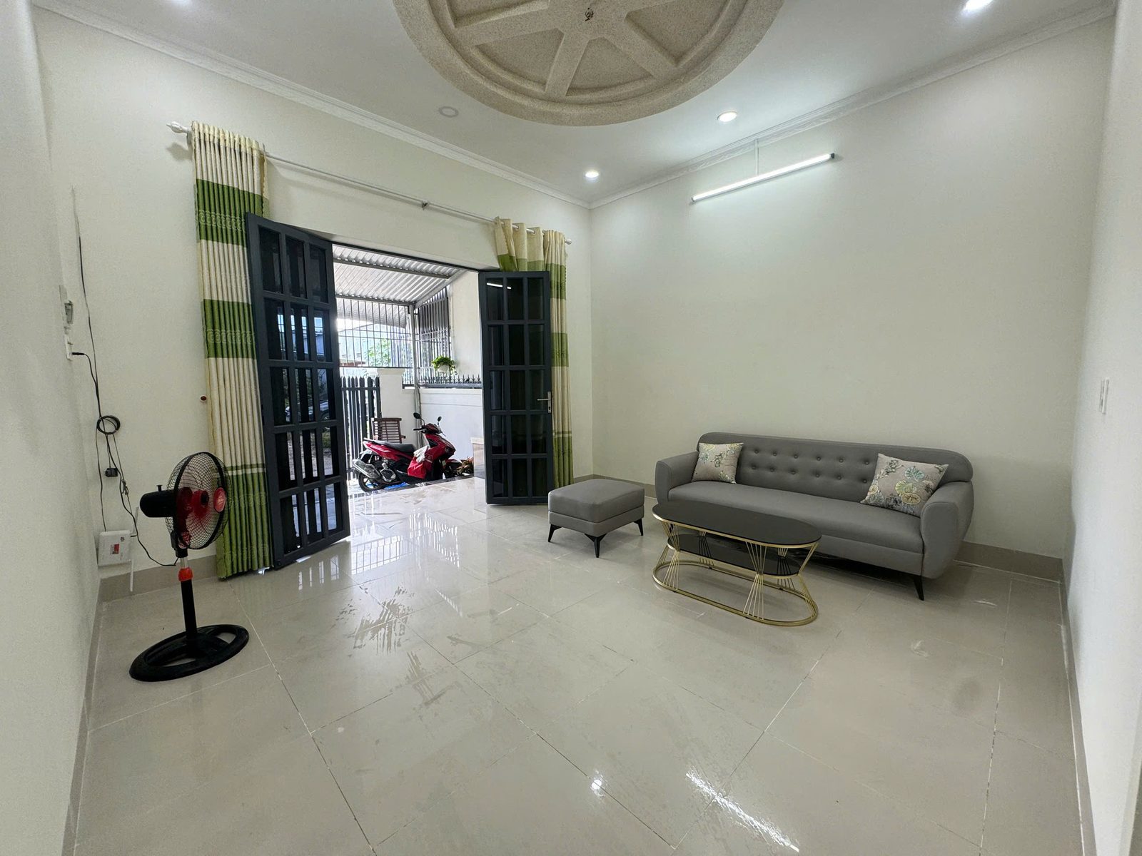 Phòng khách nhà Vĩnh Ngọc với bộ sofa xám, bàn trà, trần thạch cao mảng tròn và cửa gỗ kính mở ra sân trước