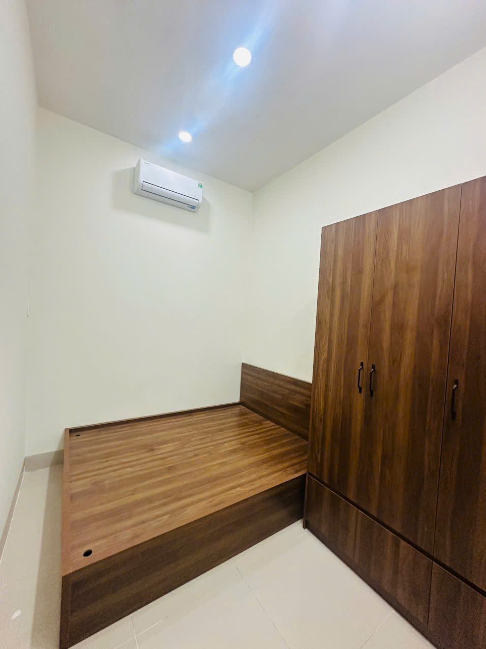Phòng ngủ với giường gỗ đóng liền bục, tủ áo ba cánh cùng tông gỗ và máy lạnh treo tường