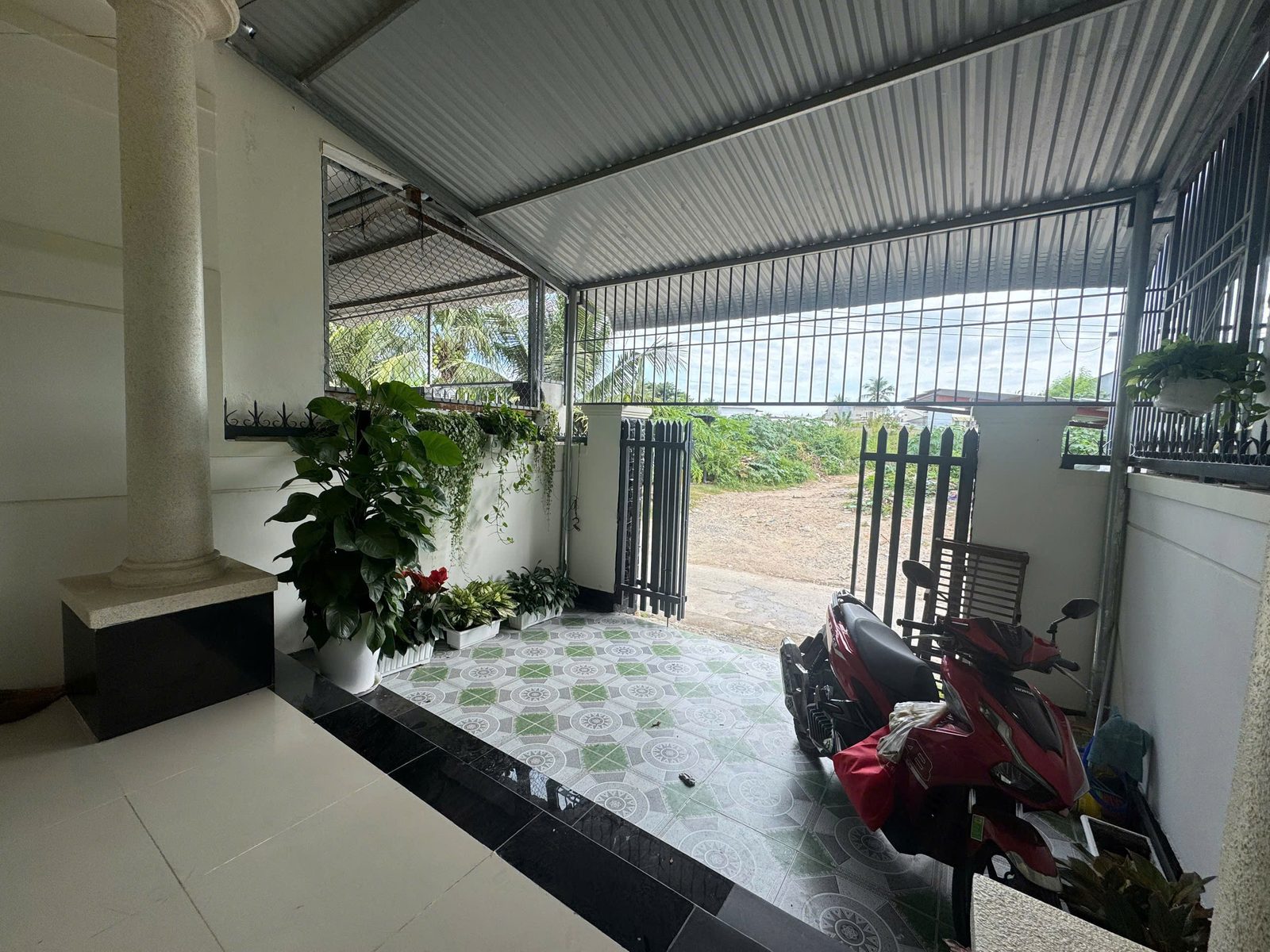 Sân trước nhà lợp mái tôn khung sắt, nền lát gạch hoa, hàng rào sắt và cổng mở ra đường trước nhà